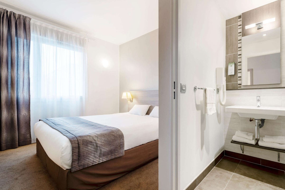 Hotellihuone, jossa on sänky, yöpöytälamppu sekä kylpyhuone, jossa on pesuallas, peili ja pyyhekoukku.