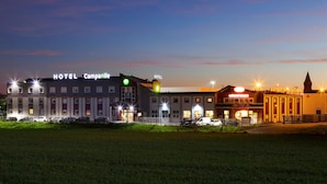 Un hotel llamado "HOTEL Campanile" con una fachada moderna, varios espacios de estacionamiento y una imponente torre del reloj al fondo.