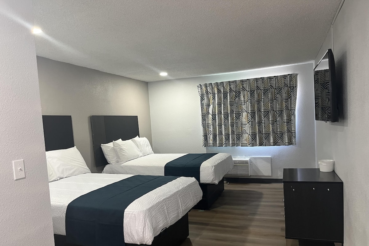 A hotel room with two beds, a flat-screen TV, a small cabinet, and a patterned curtain.
