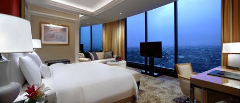 A hotel room with a large bed, a desk, a chair, a TV, and a view of the city.
