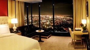 A hotel room with a large bed, a sofa, a desk, and a view of a cityscape at night. - The Trans Luxury Hotel (Bandung)