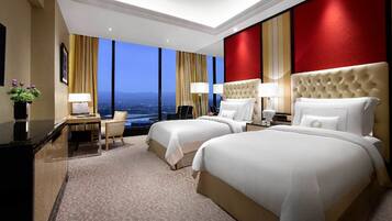 A hotel room with two beds, a desk, and a chair. The room has large windows and a view of the city.