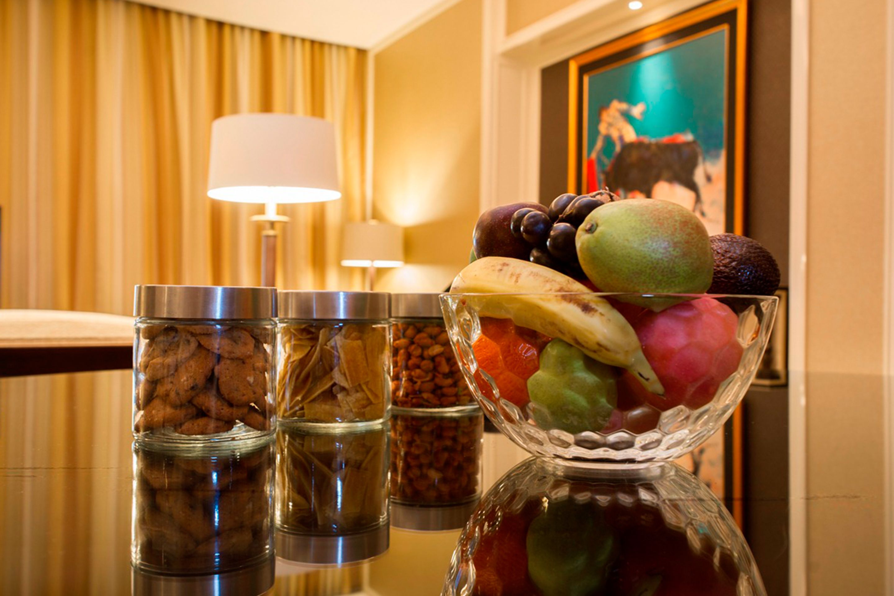 A table with a fruit bowl, jars of nuts, and a lamp in a hotel room.