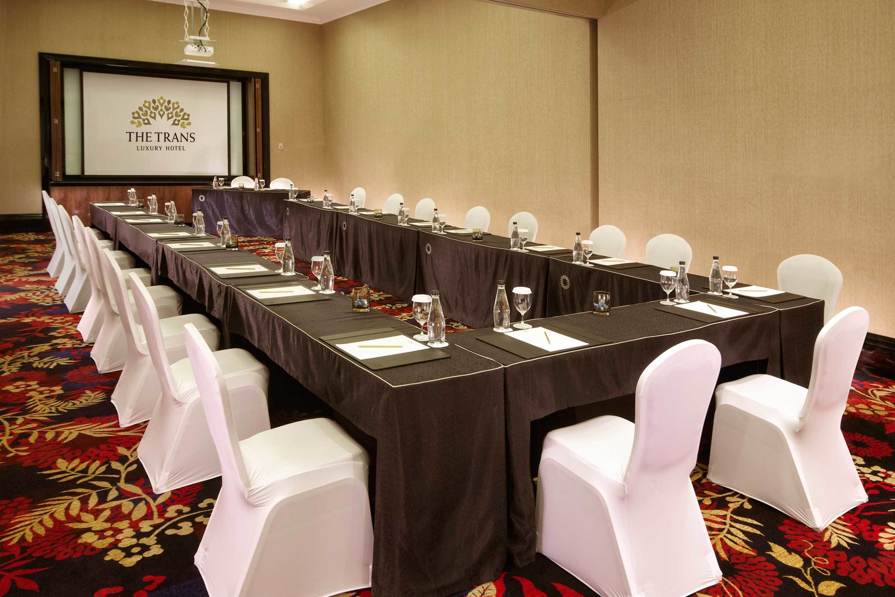 A conference room with a long table, white chairs, and a projector screen displaying "THE TRANS LUXURY HOTEL".
