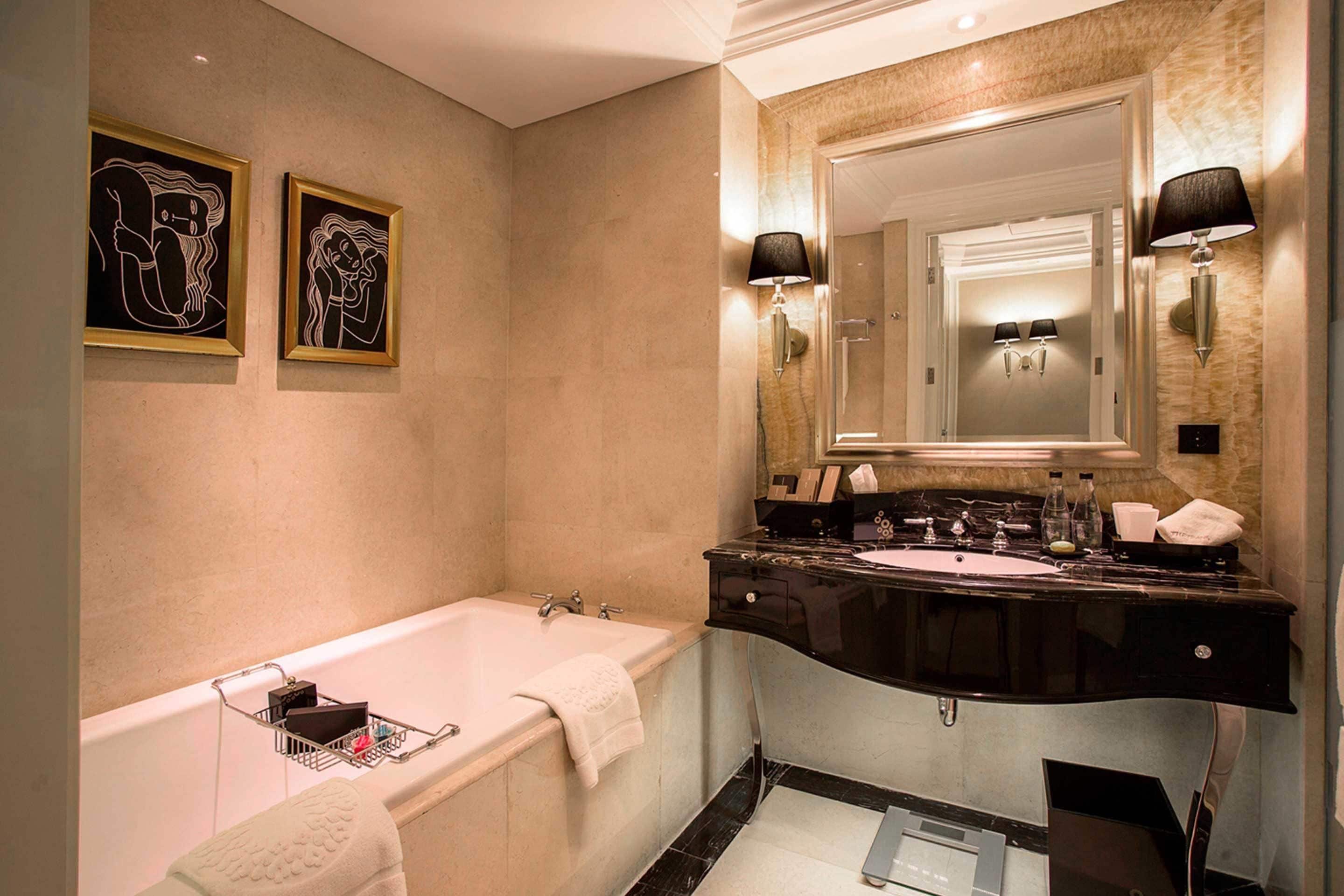 A modern bathroom with a bathtub, two framed artworks, a vanity with a mirror, and two wall-mounted lamps.