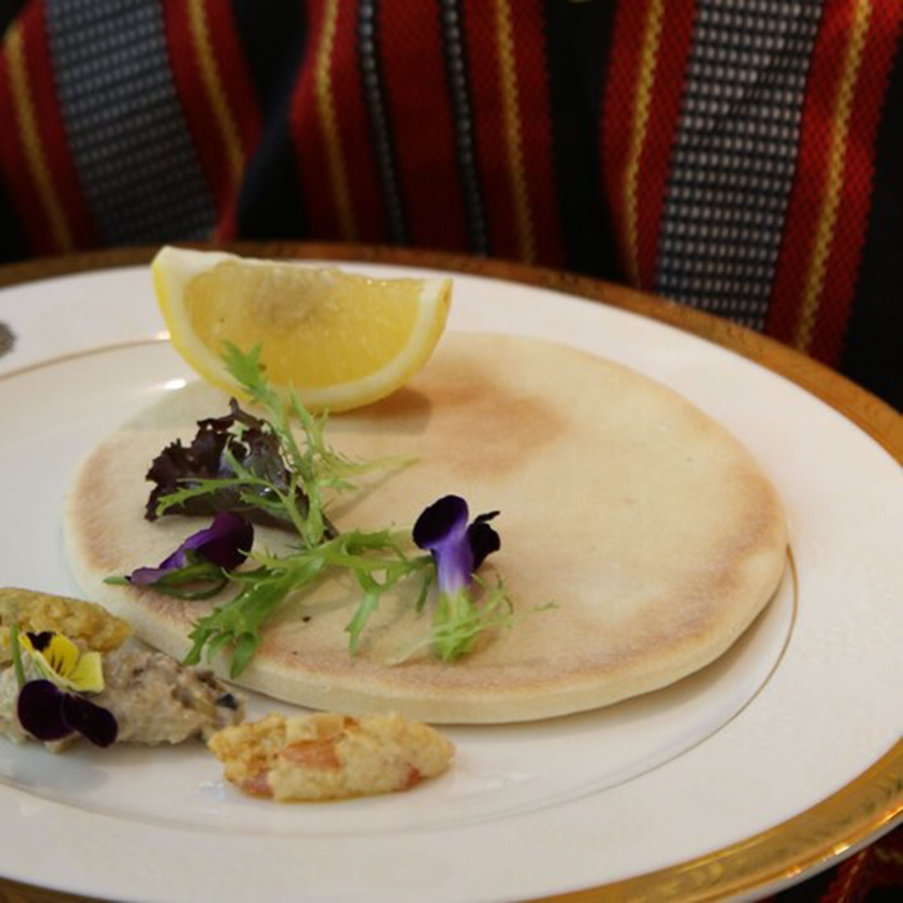 A plate with a flatbread, a lemon wedge, and some greens and a small piece of food.