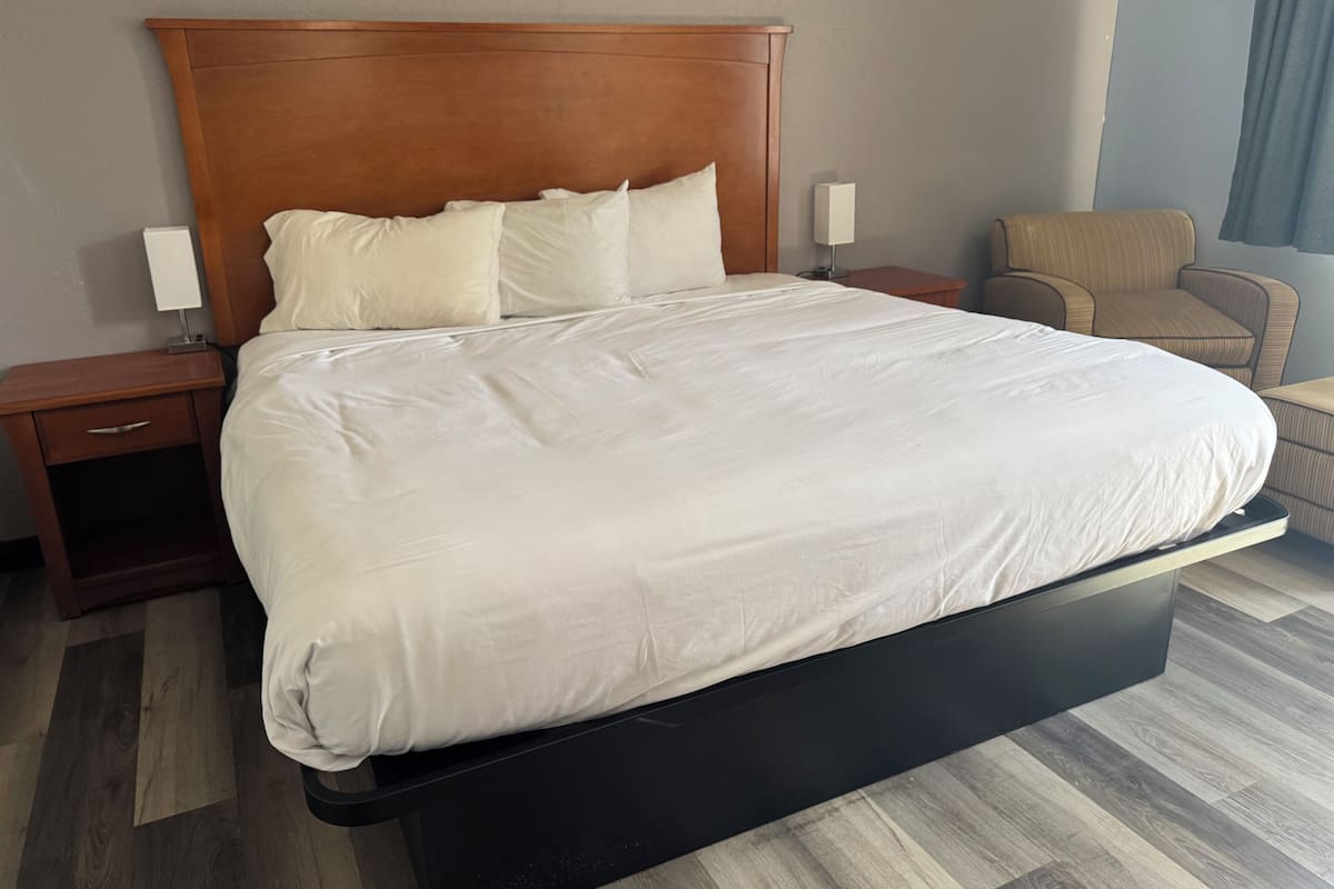 A hotel room with a large bed, a nightstand with a lamp, a chair, and a wooden floor.