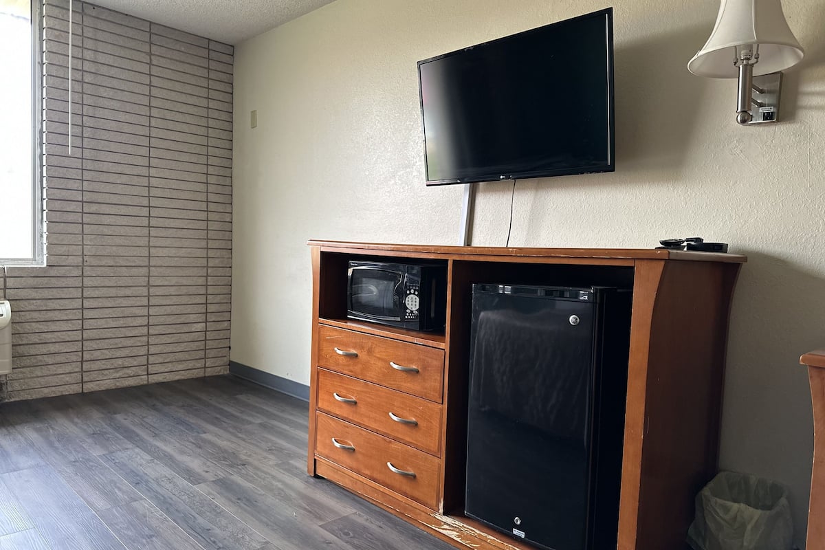 A compact room with a wooden cabinet, a microwave, a small refrigerator, a television mounted on the wall, and a window with blinds.