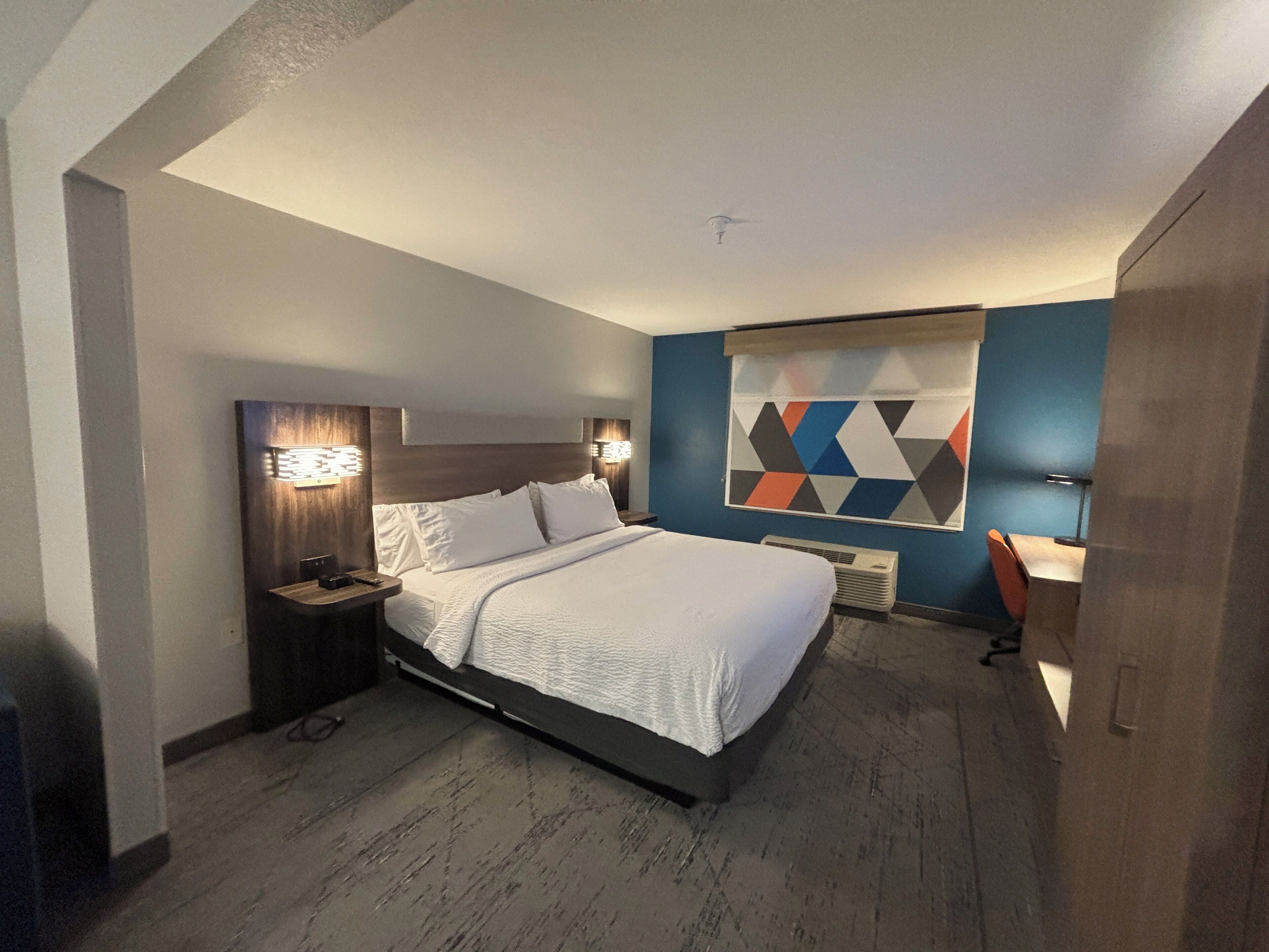 Ein modernes Hotelzimmer mit einem großen Bett, Nachttischen inklusive Lampen, einer wandmontierten Klimaanlage und einer Wanddekoration mit geometrischem Muster.