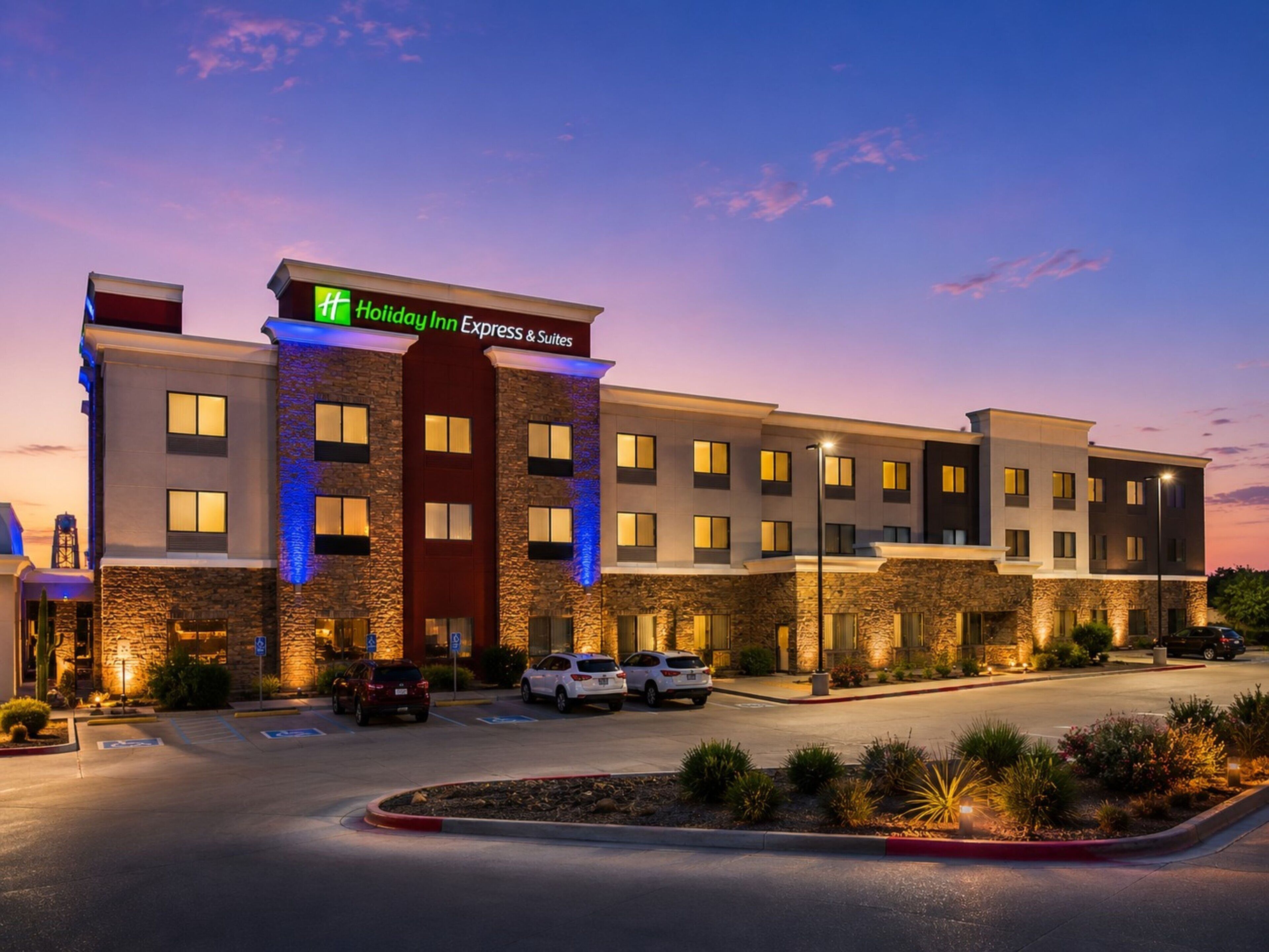 A Holiday Inn Express & Suites hotel with a modern design, featuring a stone facade, multiple parking spaces, and a landscaped entrance area.
