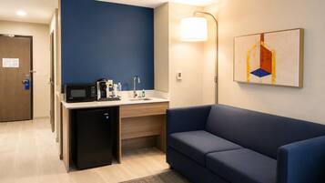 A compact hotel room with a kitchenette, a small refrigerator, a sofa, a desk, and a wall-mounted lamp.