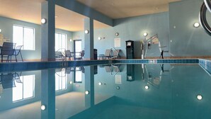 An indoor swimming pool with clear water, lounge chairs, and a poolside area with a view of the outdoors.