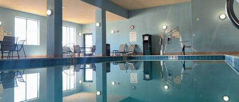 An indoor swimming pool with clear water, lounge chairs, and a poolside area with a view of the outdoors.