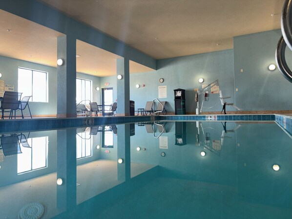 An indoor swimming pool with clear water, lounge chairs, and a poolside area with a view of the outdoors.