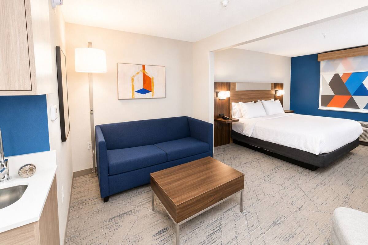 A hotel room with a bed, a sofa, a coffee table, a desk with a lamp, and a painting on the wall.