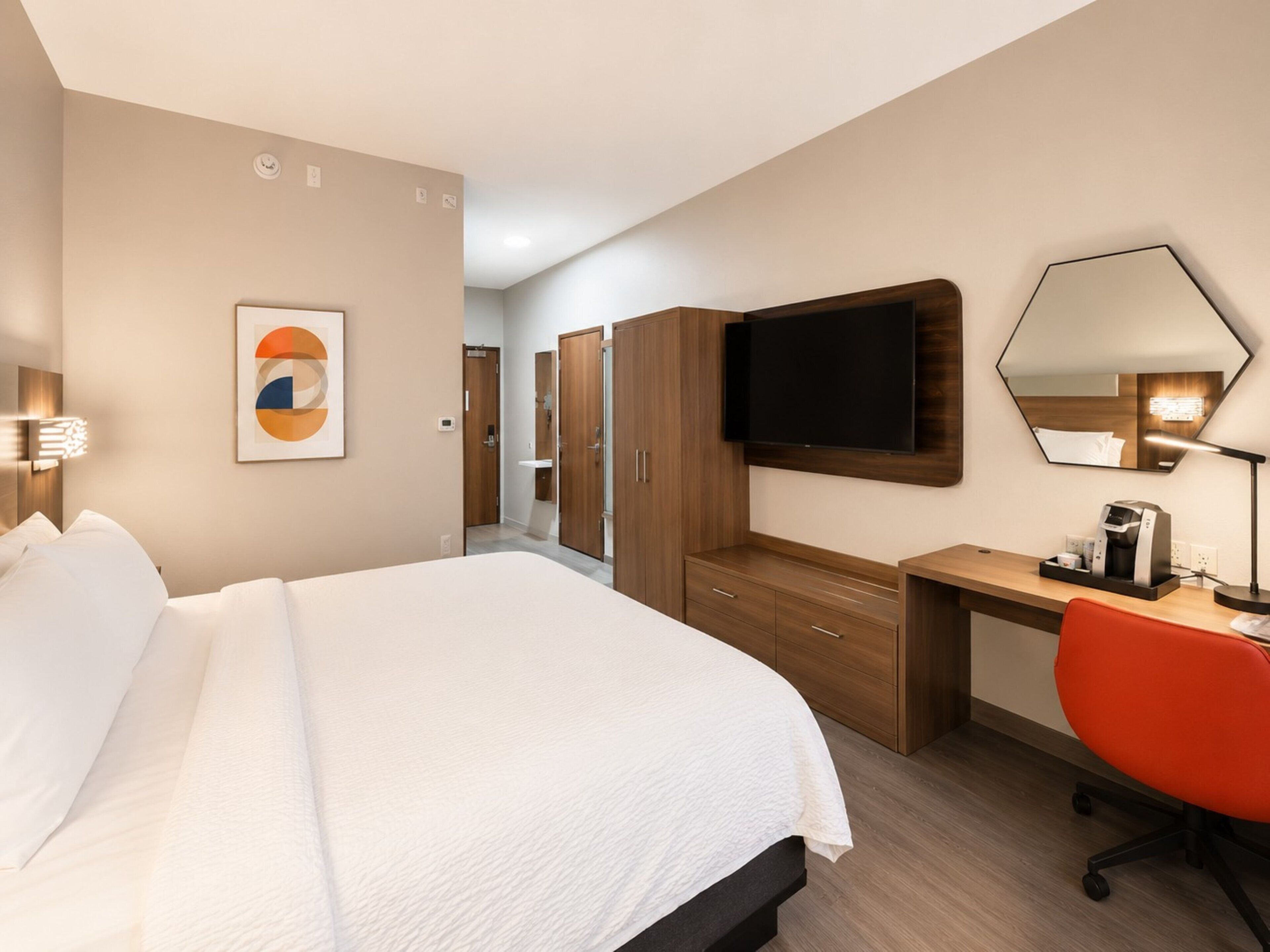 A hotel room with a bed, a desk, a chair, a TV, a mirror, and a coffee machine.