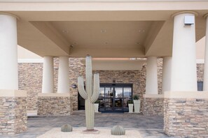 Una entrada de edificio con una gran escultura de cactus, muros de piedra y un corredor cubierto.