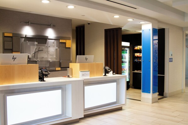 A hotel reception area with two reception desks, a digital display, and a vending machine in the background. - Holiday Inn Express and Suites Overland Park by IHG (Overland Park)