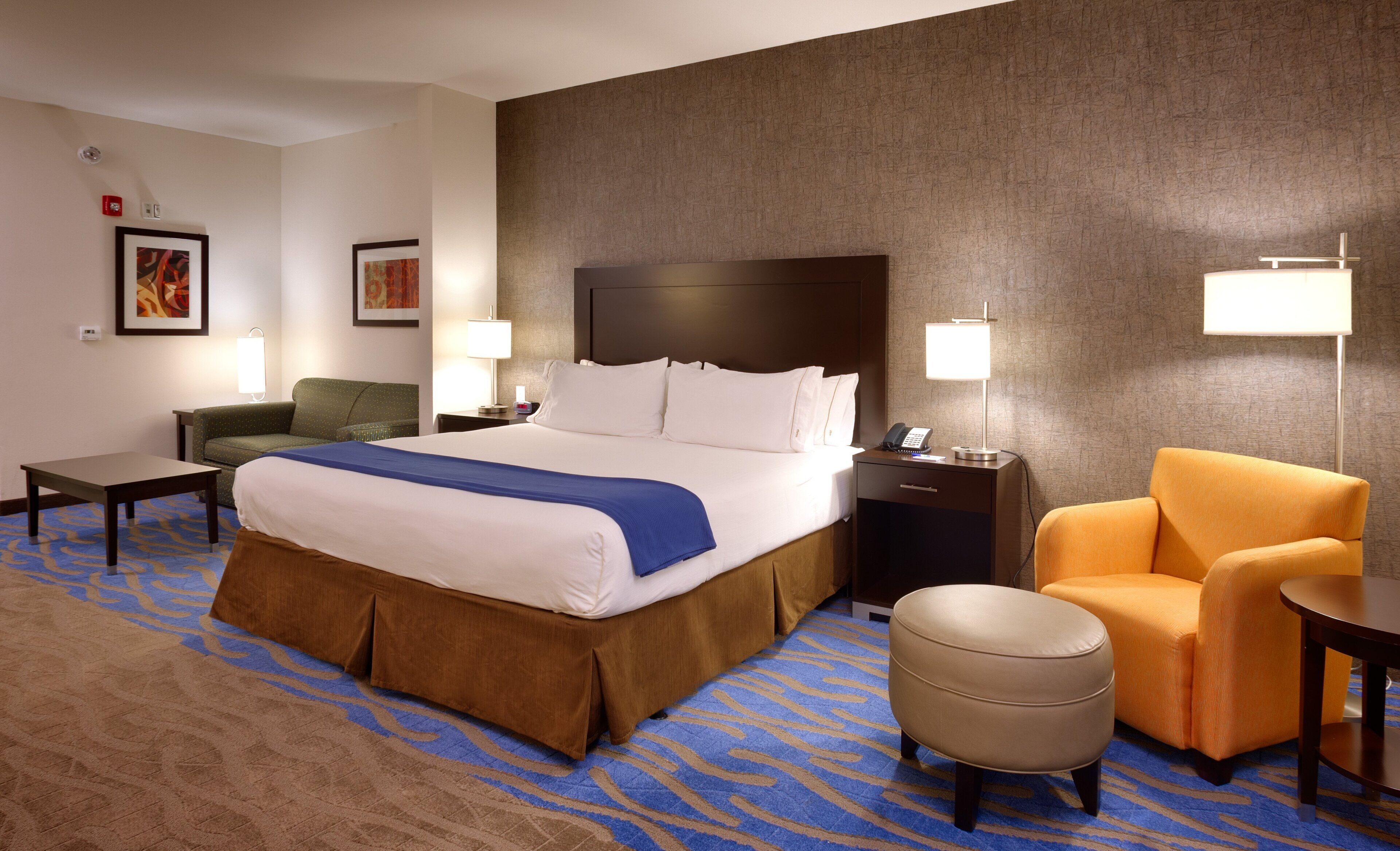 Una habitación de hotel con una cama grande, un sofá, una silla, una mesita y una mesita de noche con una lámpara.
