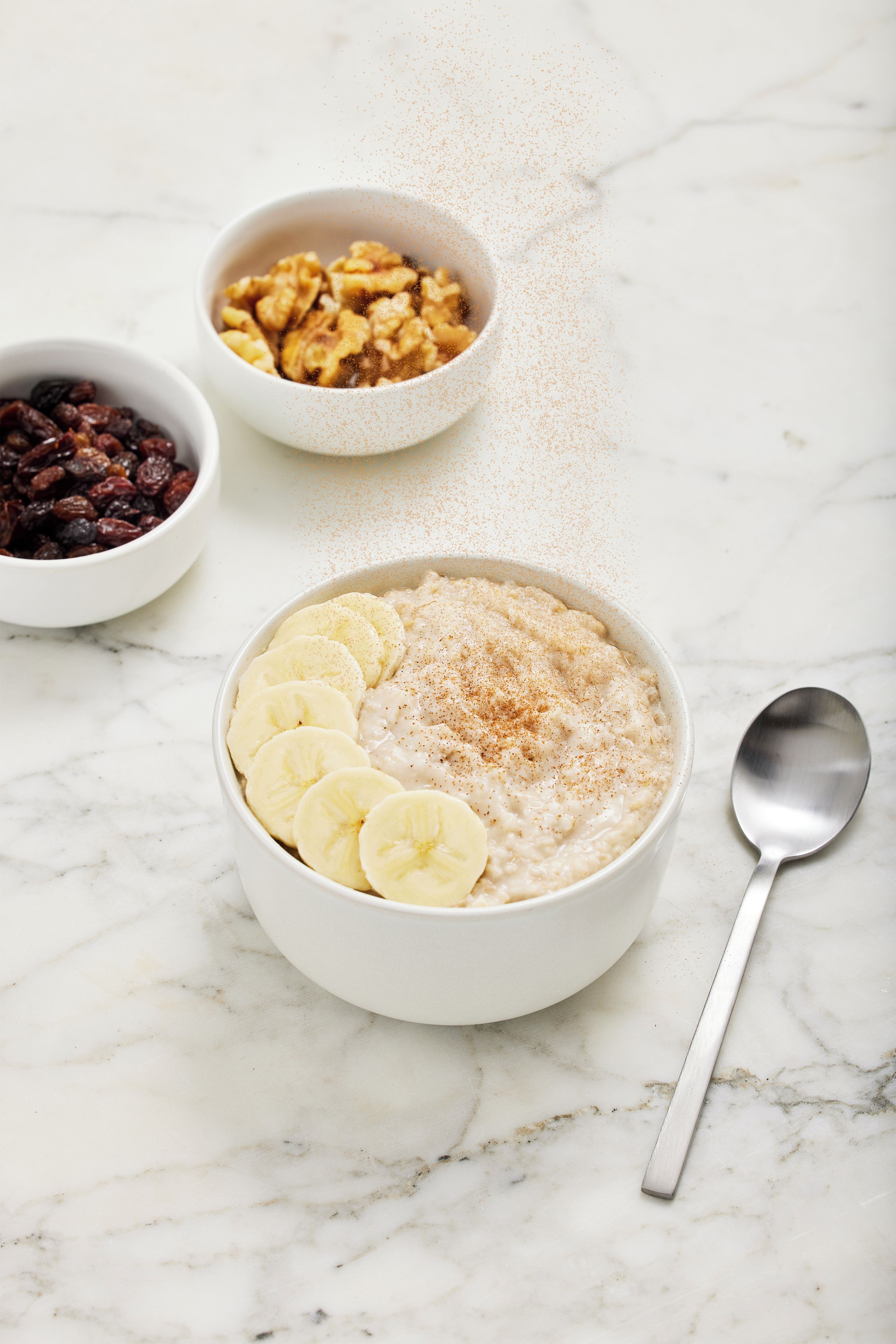 A bowl of oatmeal with banana slices and cinnamon, a bowl of dried fruits, and a bowl of granola on a marble surface.