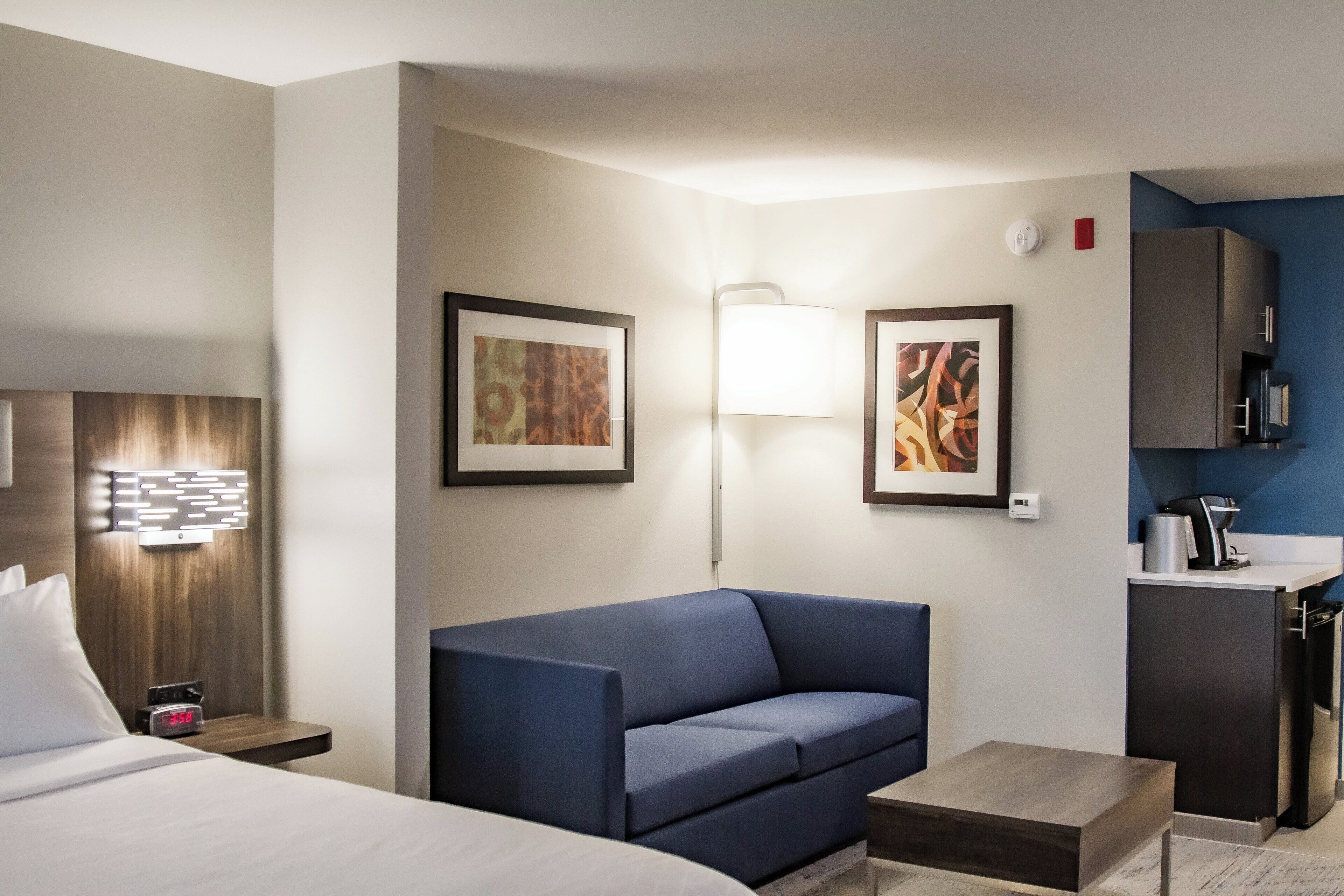 Quarto de hotel com cama, sofá azul, mesinha, escrivaninha, abajur e quadros nas paredes.