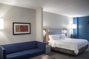 A hotel room with a bed, a blue sofa, a framed picture on the wall, and a bedside table with a lamp. - Holiday Inn Express and Suites Overland Park by IHG (Overland Park)
