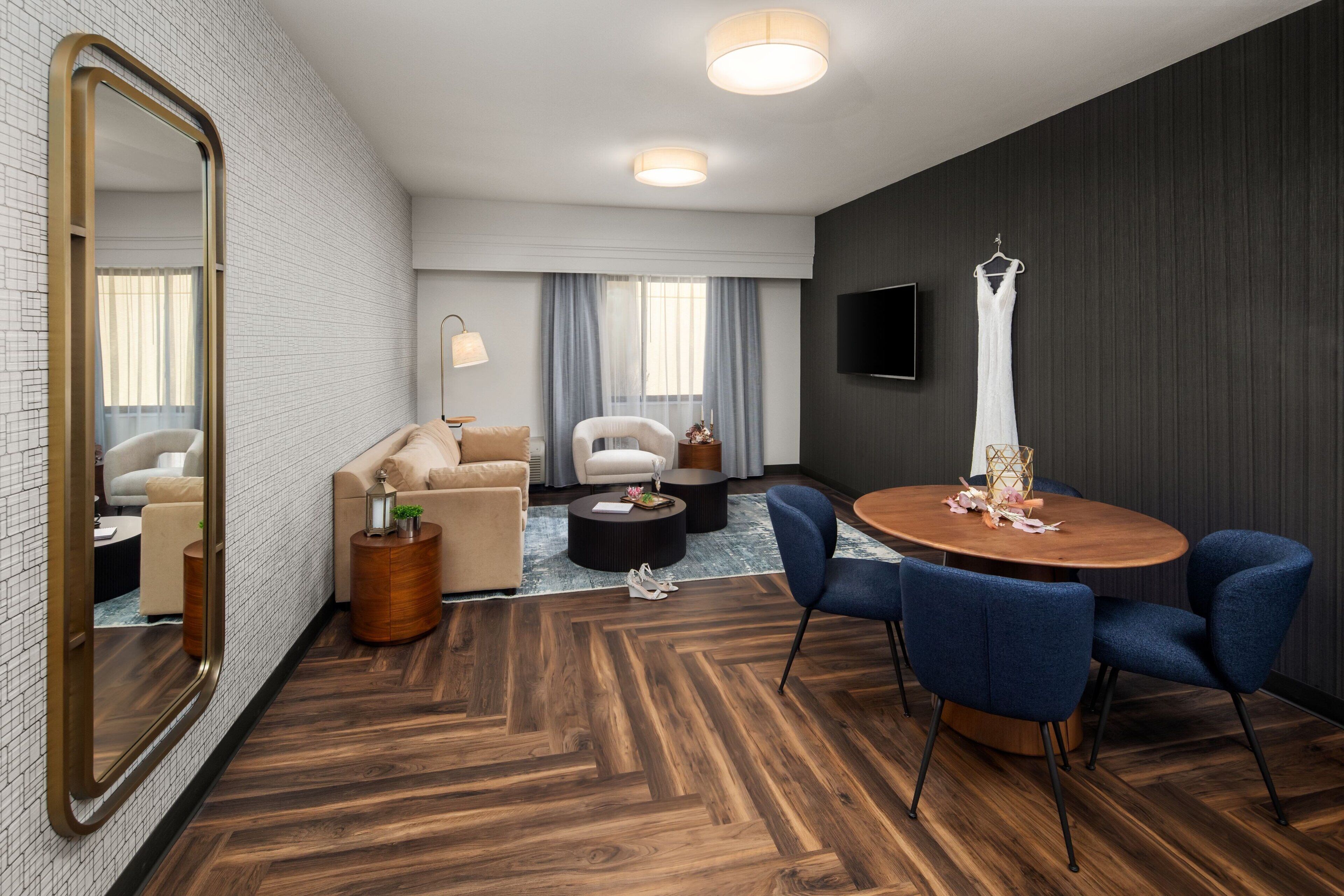 A modern hotel room with a dining area, a large mirror, a TV, and a hanging dress.