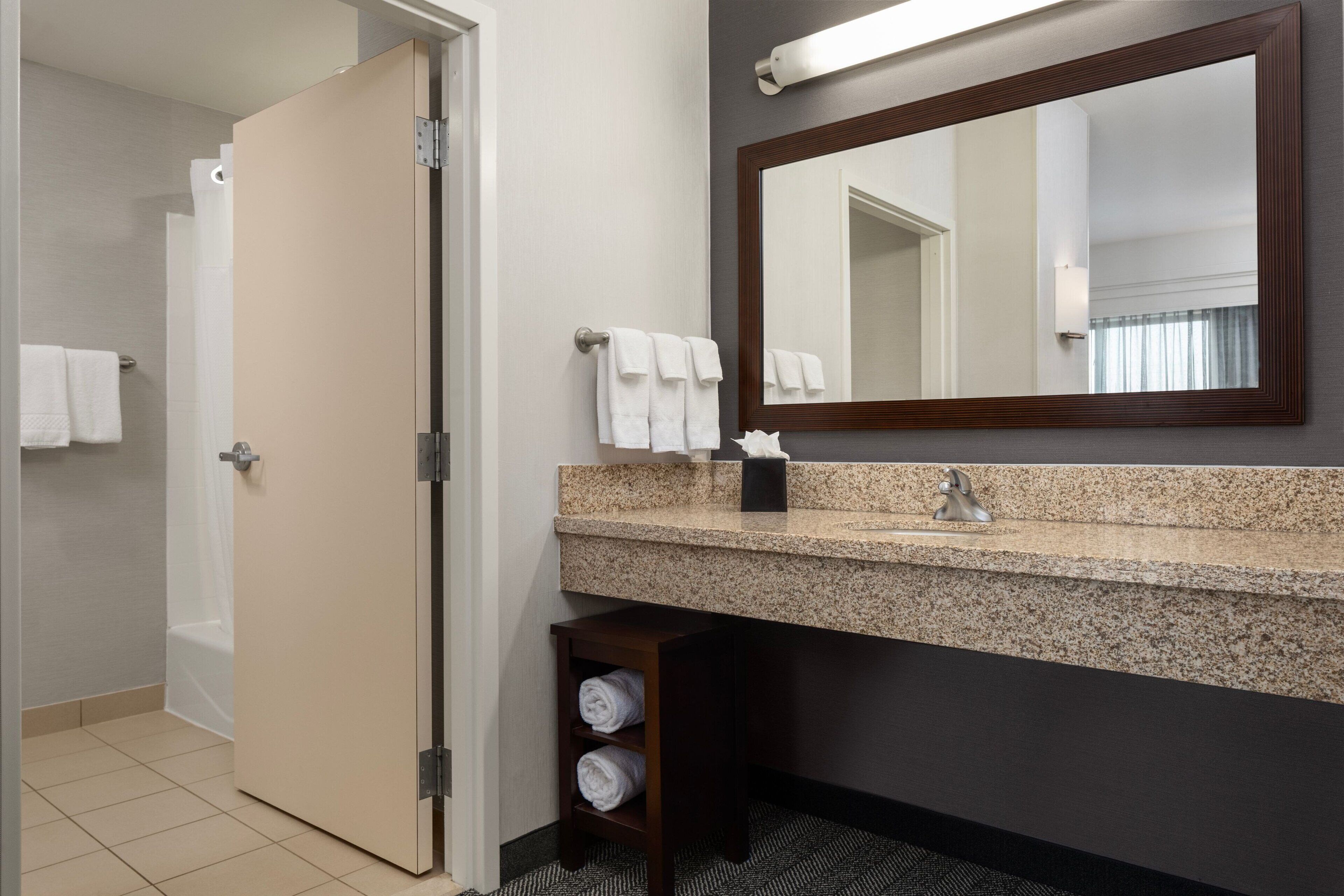 A bathroom with a granite countertop, a mirror, a towel rack, and a small shelf with towels.