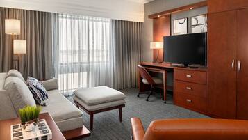 A hotel room with a sofa, ottoman, desk, television, and a cabinet.