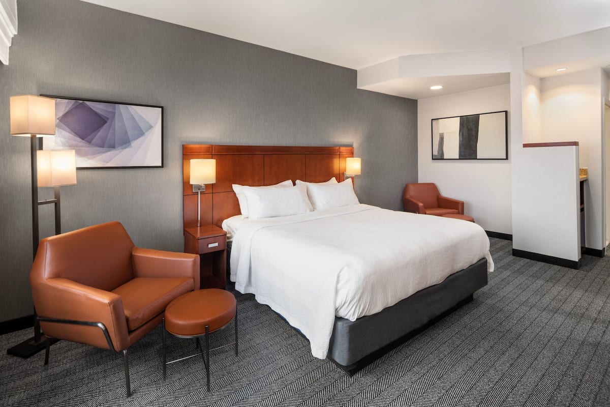 A hotel room with a large bed, two bedside tables with lamps, a brown leather chair, a small round table, and a wall-mounted artwork.