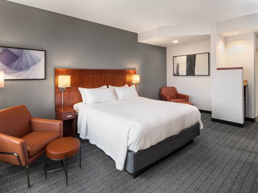 Een hotelkamer met een groot bed, twee nachtkastjes met lampen, een bruine leren fauteuil, een klein rond tafeltje en een kunstwerk aan de muur.
