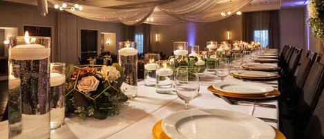 A banquet hall with long tables set for a formal event, featuring white tablecloths, gold-rimmed plates, and candles.