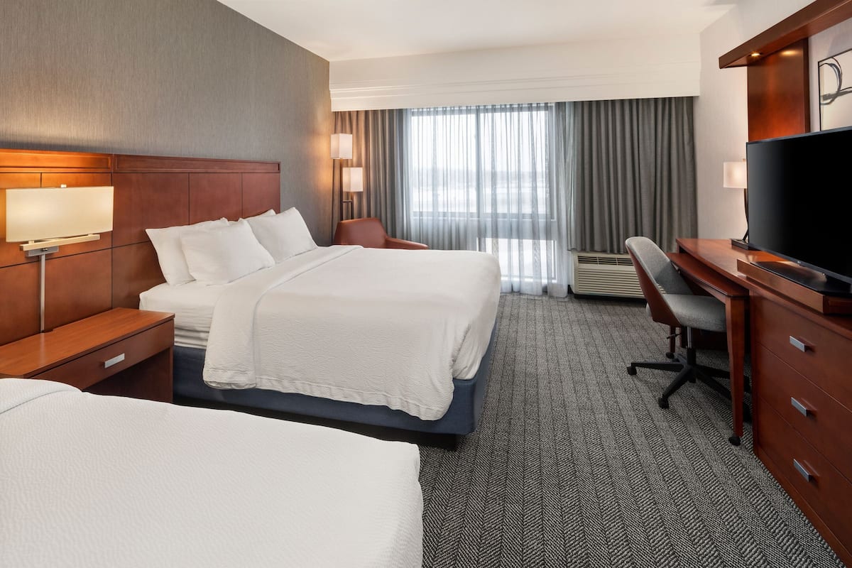 A hotel room with two beds, a desk, a television, and a large window with curtains.