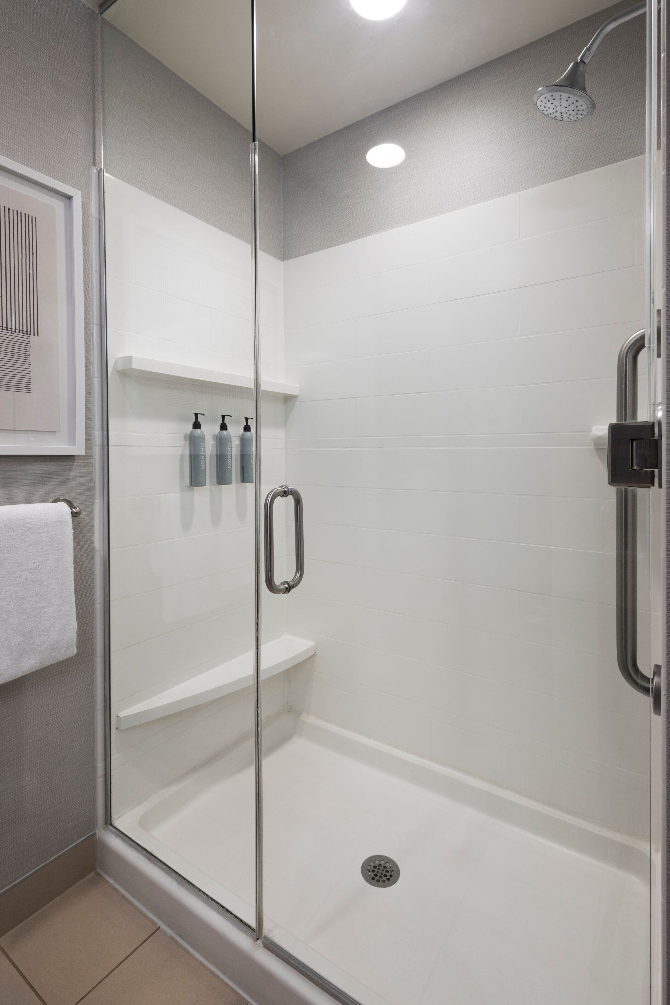 A glass-enclosed shower stall with a handheld showerhead, a white shelf with toiletries, and a white towel on the floor.