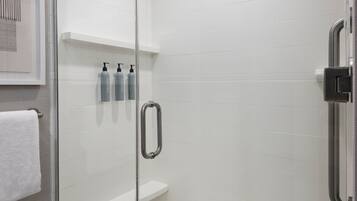 A glass-enclosed shower stall with a handheld showerhead, a white shelf with toiletries, and a white towel on the floor.