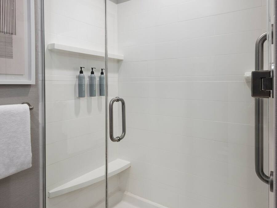A glass-enclosed shower stall with a handheld showerhead, a white shelf with toiletries, and a white towel on the floor.