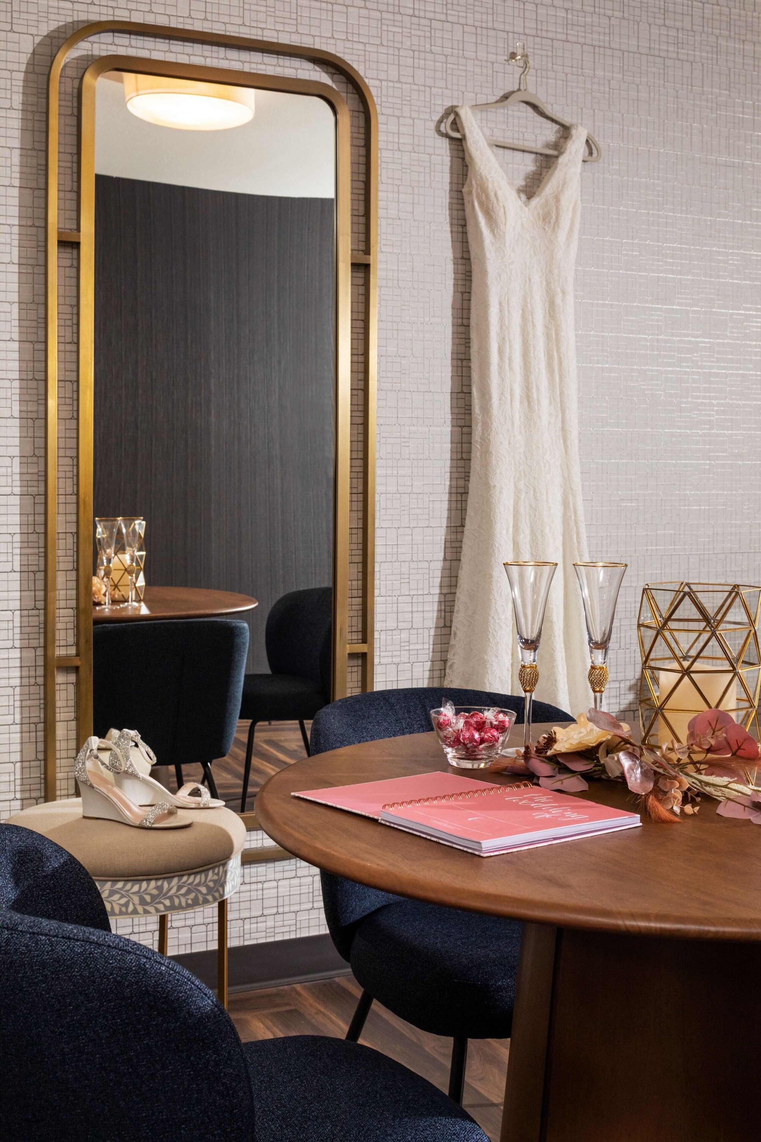 Um quarto com uma mesa redonda de madeira, duas cadeiras, um espelho grande, um vestido pendurado e uma pequena mesa com um vaso e um caderno.