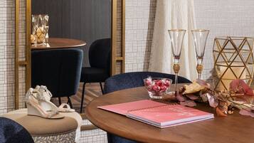 Um quarto com uma mesa redonda de madeira, duas cadeiras, um espelho grande, um vestido pendurado e uma pequena mesa com um vaso e um caderno.