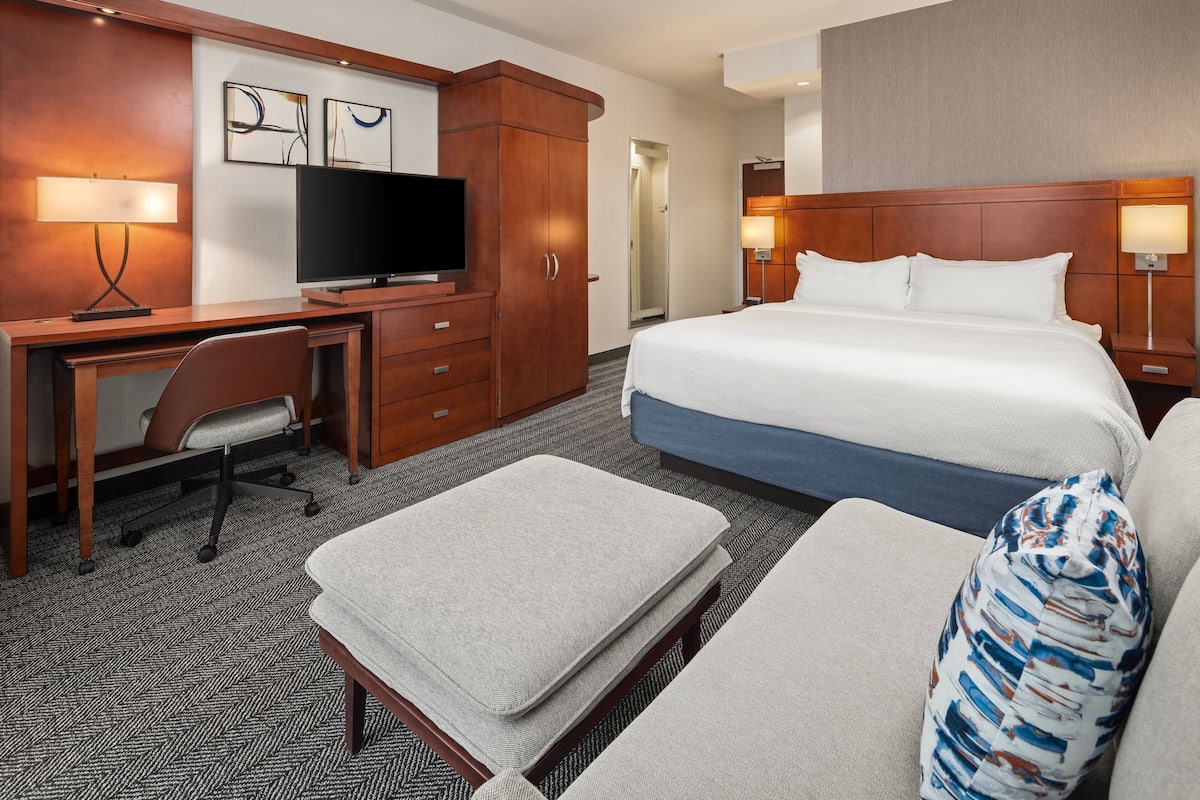 A hotel room with a bed, a desk with a television, a chair, a sofa, and a small table.