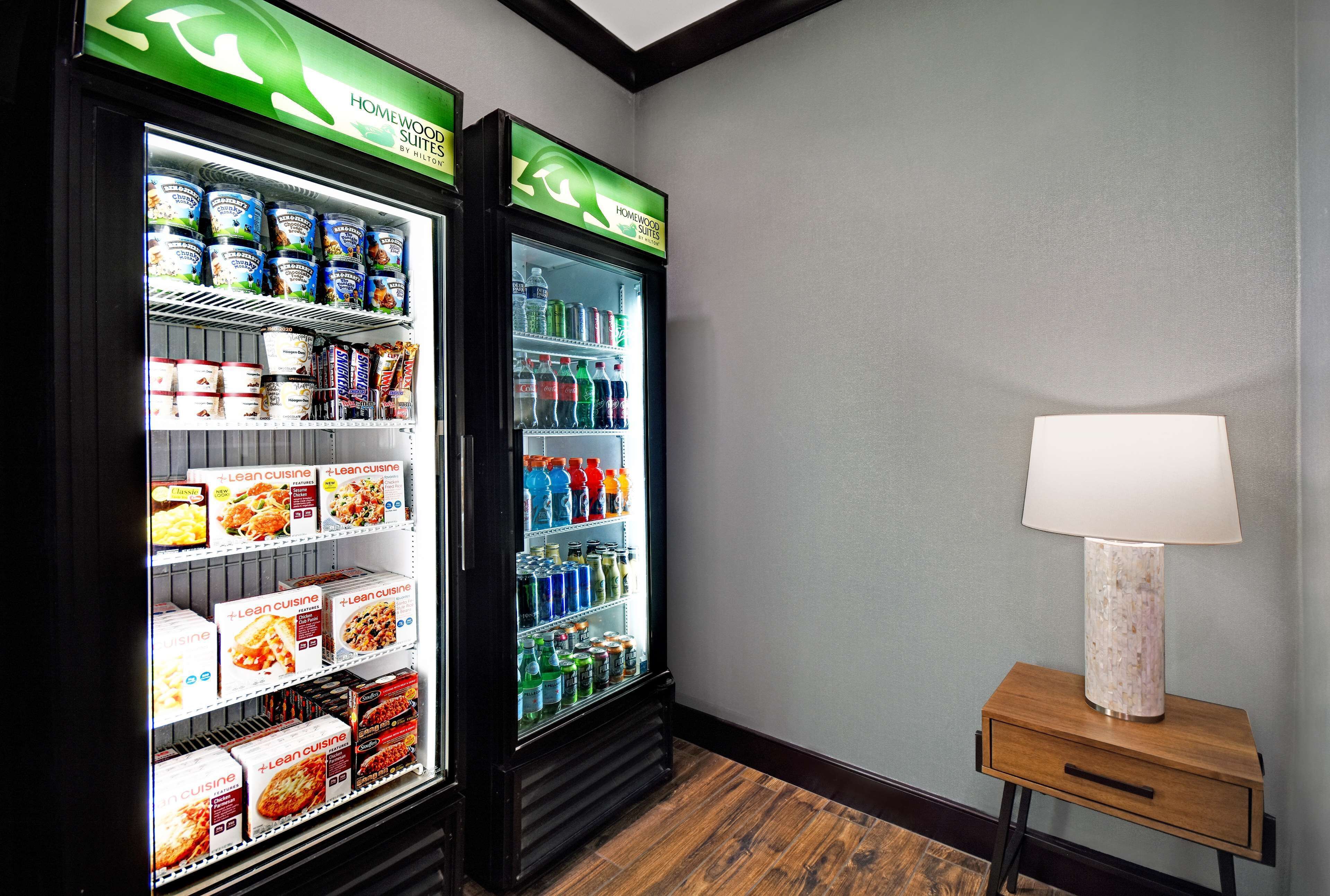 Habitación de hotel con dos vitrinas refrigeradas llenas de comida y bebidas, una mesita de noche con lámpara, y piso de madera.