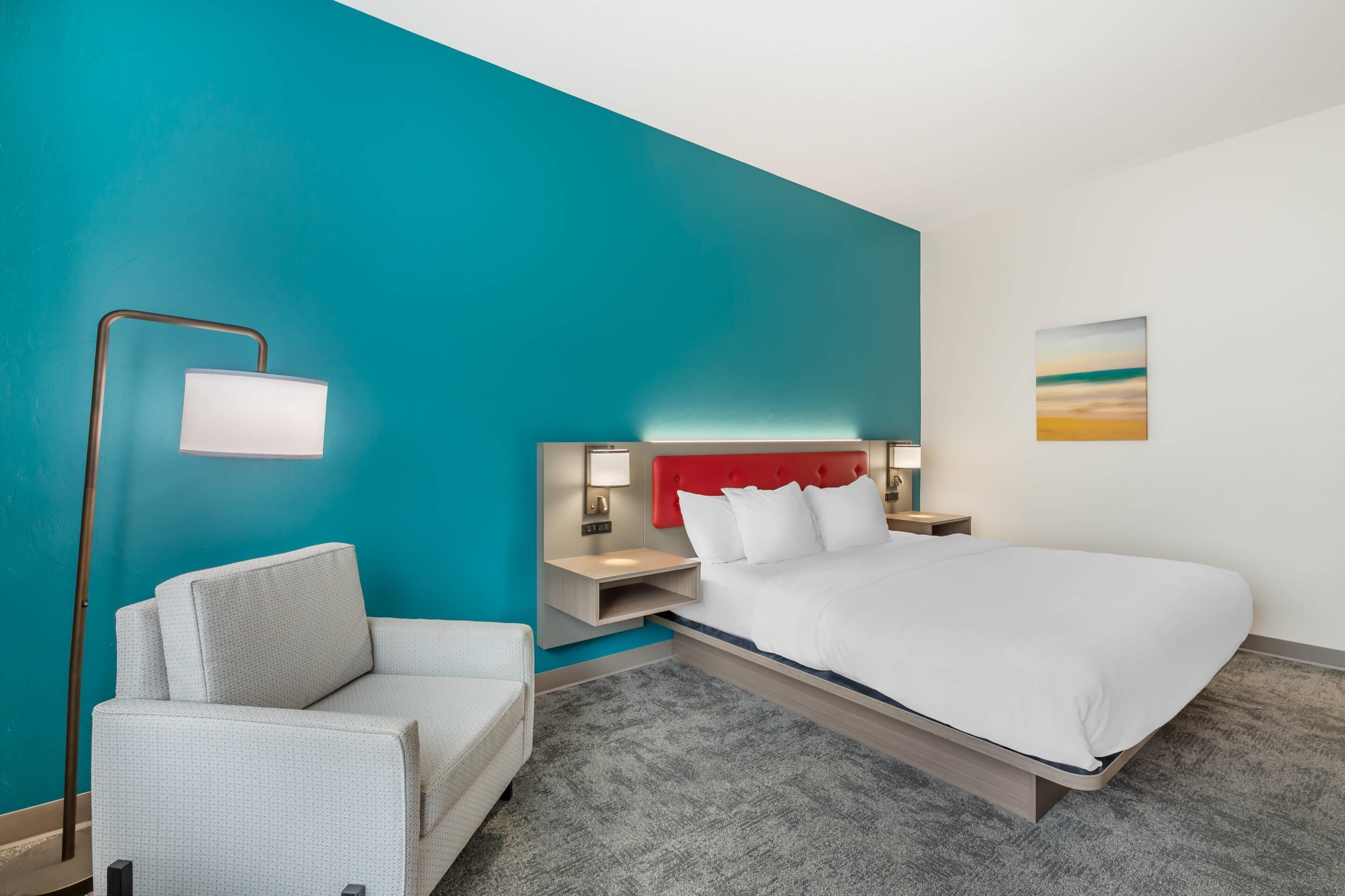 A hotel room with a bed, a chair, a nightstand, a lamp, and a painting on the wall.