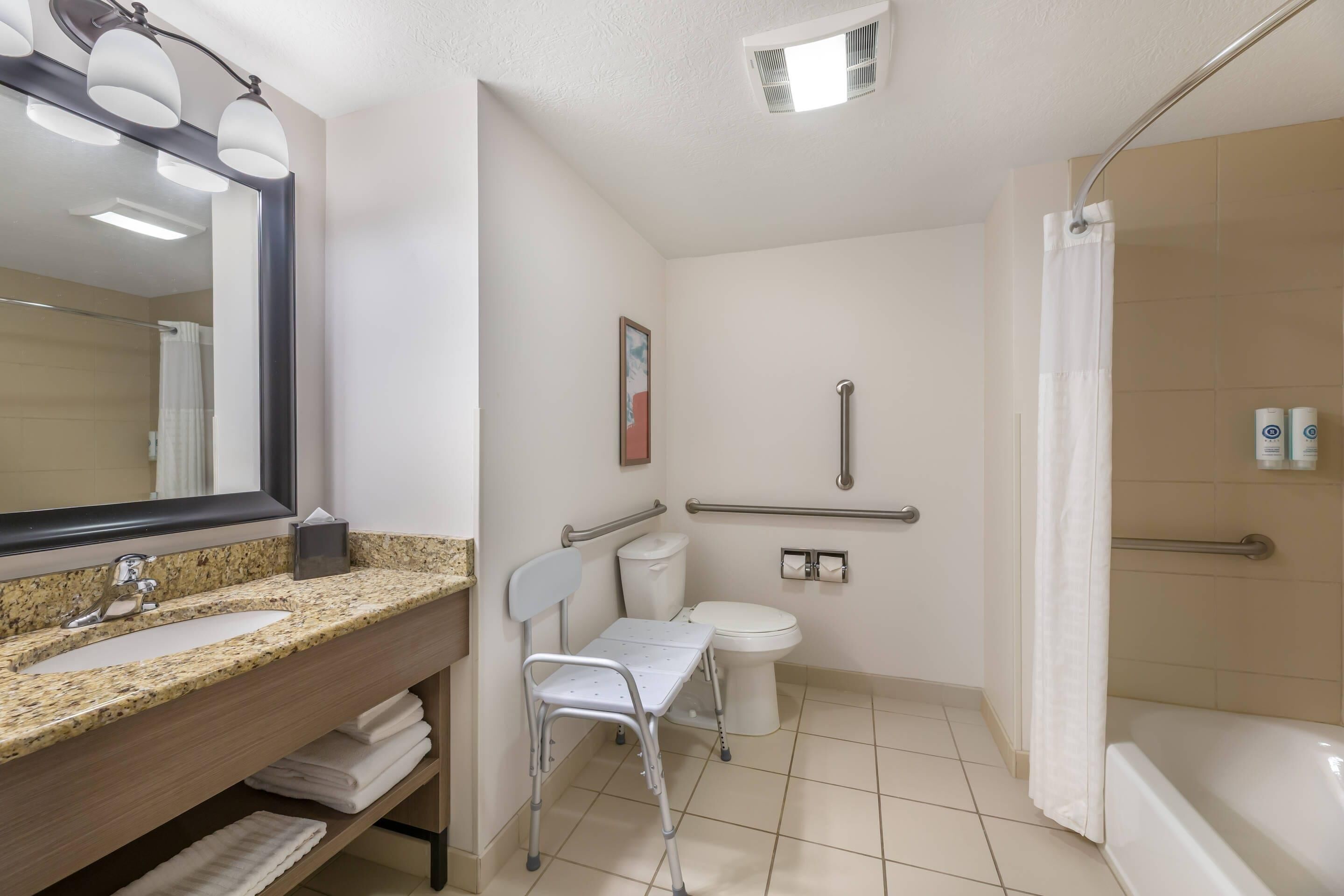 A bathroom with a granite sink, a mirror, a toilet, a shower, and a wheelchair-accessible chair.