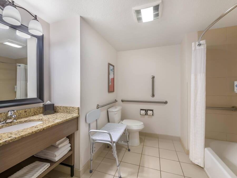 A bathroom with a granite sink, a mirror, a toilet, a shower, and a wheelchair-accessible chair.