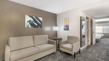 A hotel room with a sofa, armchair, a small table with a lamp, and a bed.