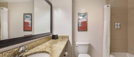 A bathroom with a granite countertop sink, a white toilet, a shower with a white curtain, and two framed abstract paintings on the wall.