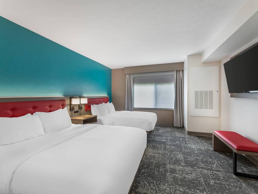 A hotel room with two beds, a red headboard, a flat-screen TV, a bench, and a window with curtains.