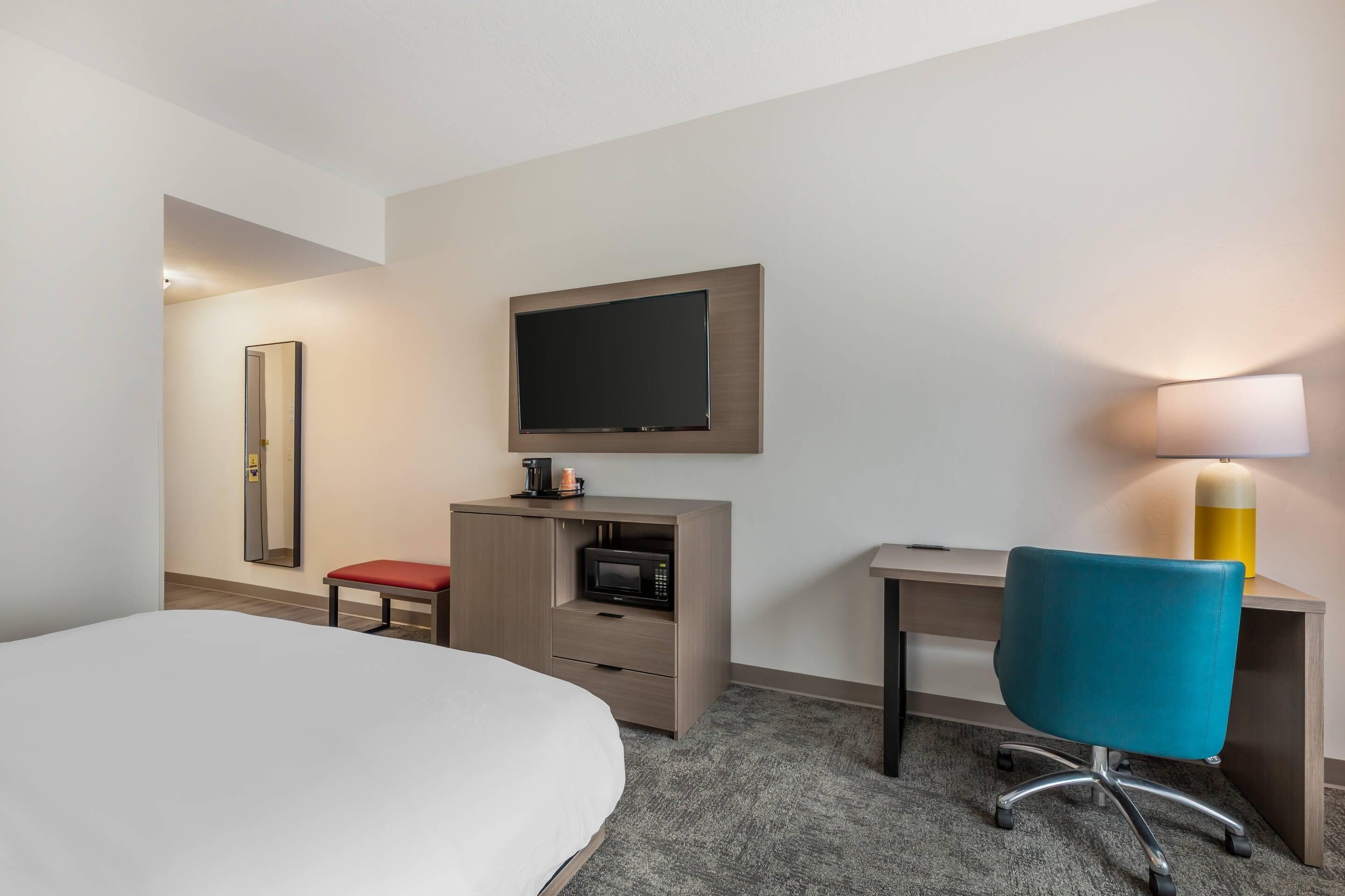 A hotel room with a bed, a desk, a chair, a television, a microwave, a lamp, and a mirror.