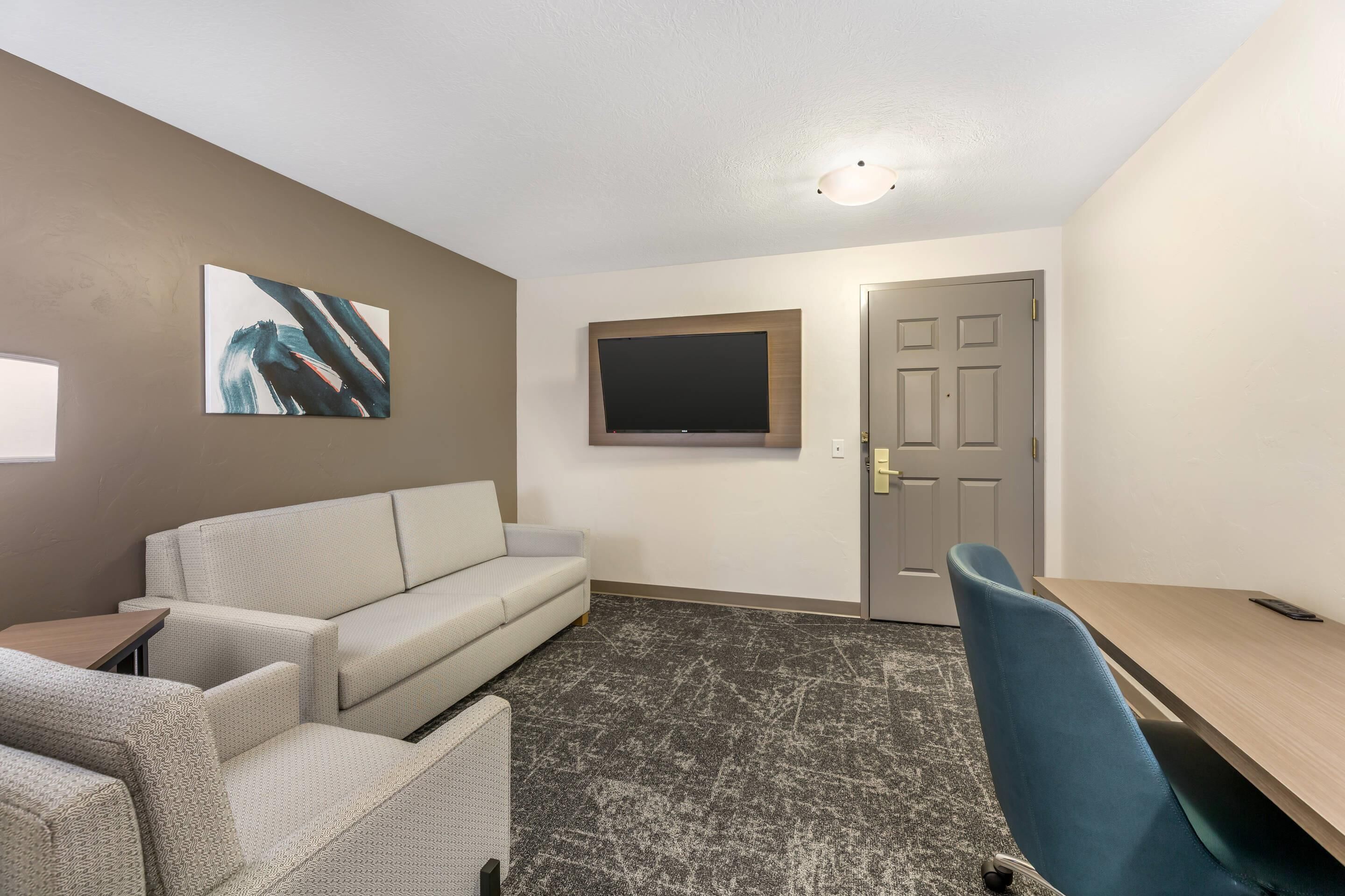 Une pièce comprenant un canapé, une petite table, un téléviseur fixé au mur, une porte et un sol recouvert d’une moquette.