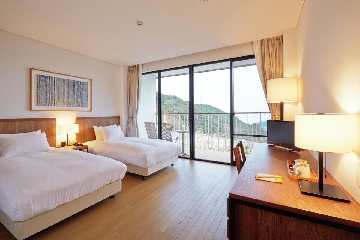 Ein Hotelzimmer mit zwei Betten, einem Schreibtisch mit Lampe, einem Fernseher und einem Balkon mit Blick auf die Hügel.