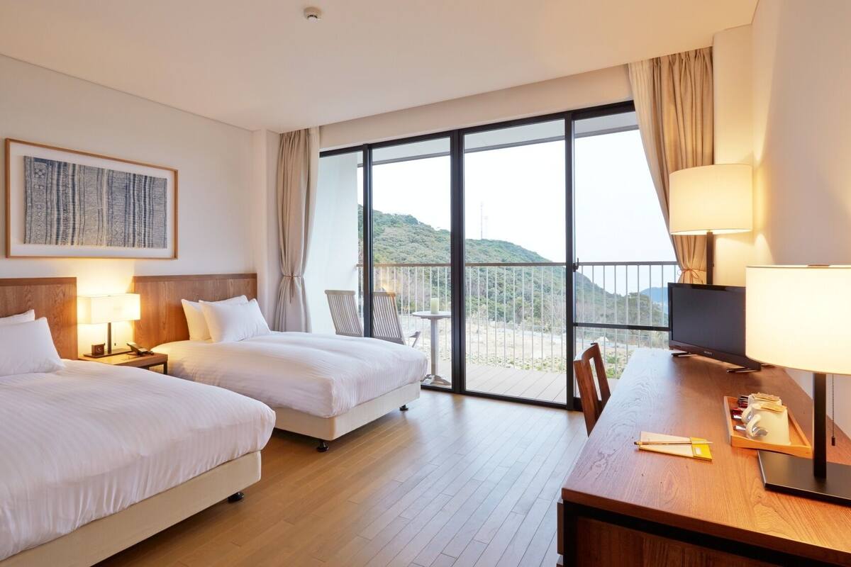 Ein Hotelzimmer mit zwei Betten, einem Schreibtisch, einem Fernseher und einem Balkon mit Blick auf die Hügel.